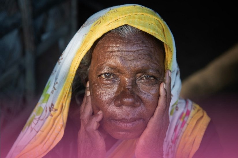 A close-up portrait of an elderly woman with her hands resting against her cheeks, looking directly at the camera. Overlaid on the image is a quote in white text: "There is a crack in everything, that's how the light gets in" attributed to Sivananthi Thanenthiran. A pink gradient fades up from the bottom.